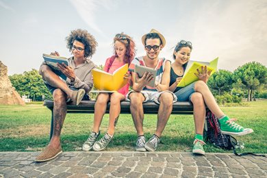 four college students sitting on a bench on campus with notebooks and a tablet