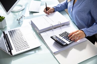 person at desk with laptop, calculator and binder of invoices